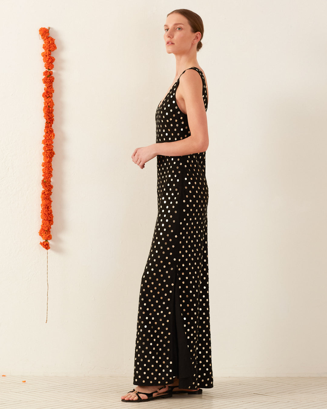 Wearing the Adele Night Sky Coin Dress, a long, sleeveless black silk slip dress adorned with white polka dots, the person stands in profile against a plain white wall. An orange marigold garland hangs vertically on the wall. They complement the outfit with black sandals and have their hair tied back.