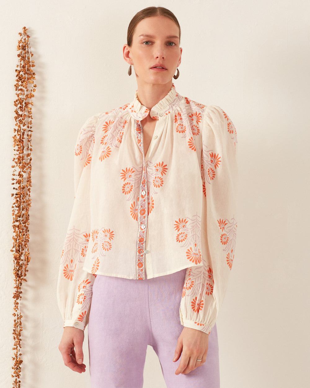 A person stands against a white background, wearing the Ana Papaya Aster Shirt, which is adorned with hand-beaded orange floral patterns and paired with light purple pants. The shirt features long sleeves and buttons down the front. They have a neutral expression and slender chain earrings in each ear.