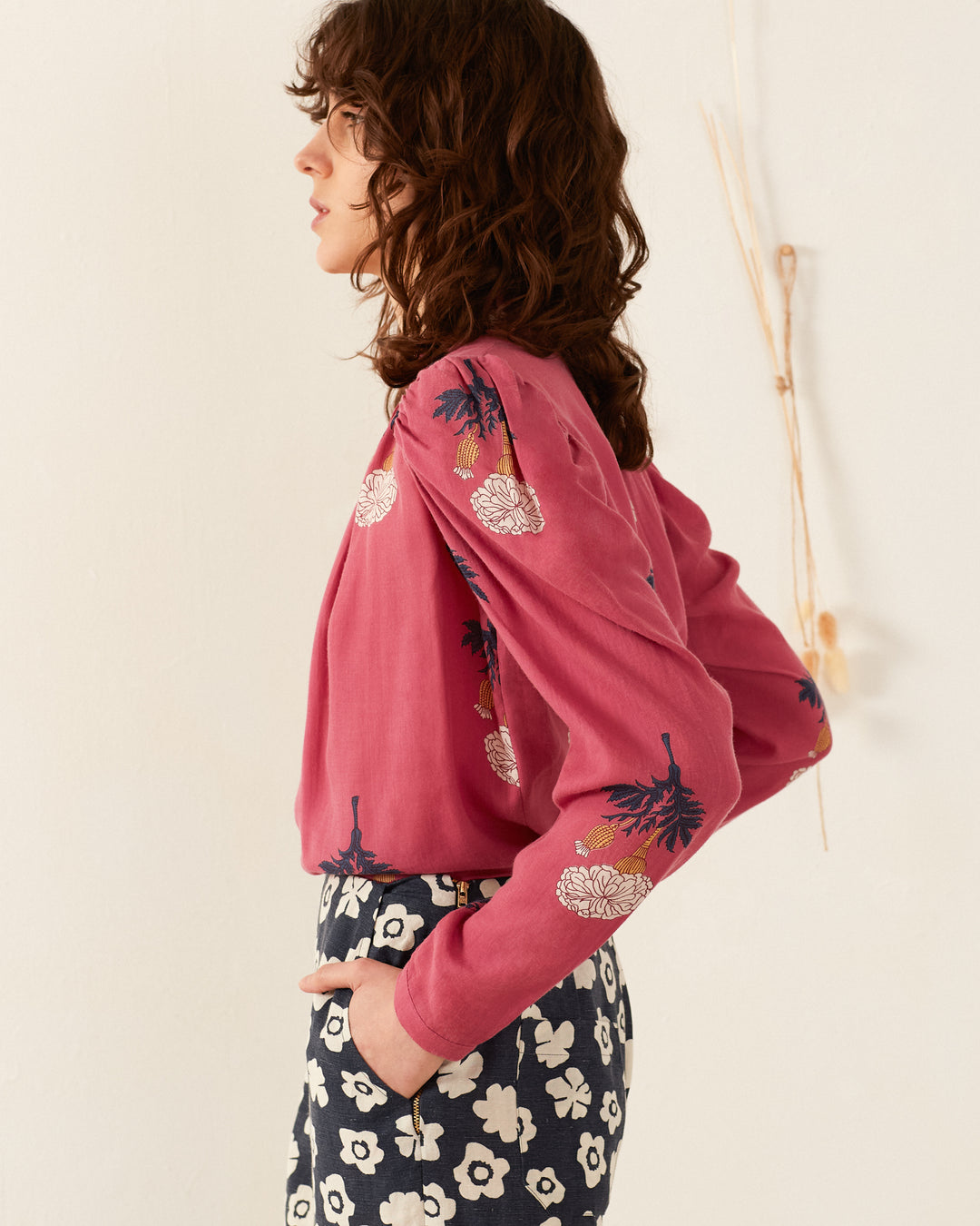 A person with wavy brown hair stands sideways, wearing the Arte Hibiscus Rose Shirt, which features a pink hue and floral embroidery on the sleeves. They pair it with high-waisted pants in a black and white floral pattern, creating an ensemble with a vintage feel against the neutral backdrop.