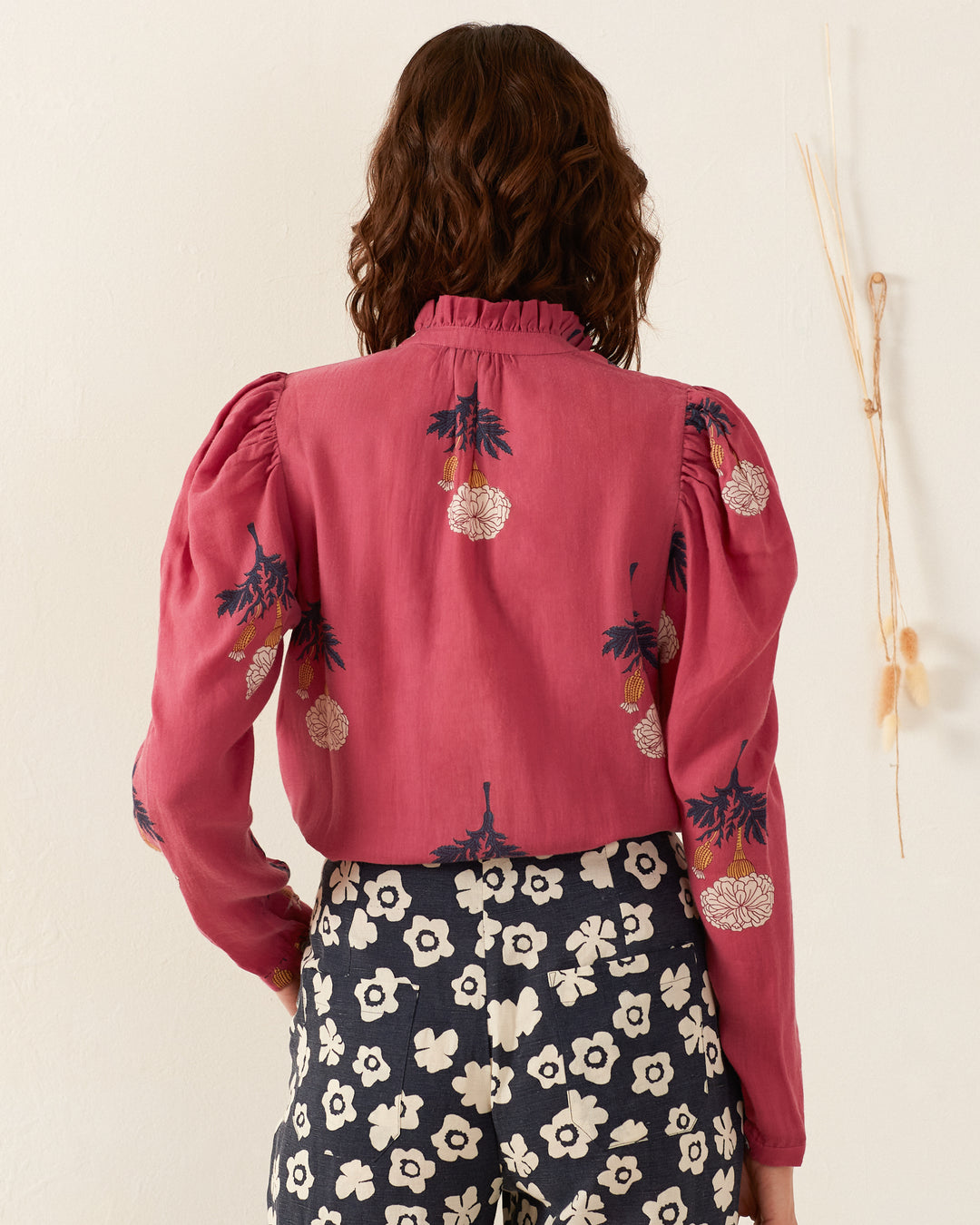 A person with long hair stylishly wears the Arte Hibiscus Rose Shirt, featuring hand-embroidered floral designs and puffy sleeves. They pair it elegantly with black and white floral patterned pants, standing against a neutral backdrop accented by decorative dried branches for a vintage ambiance.