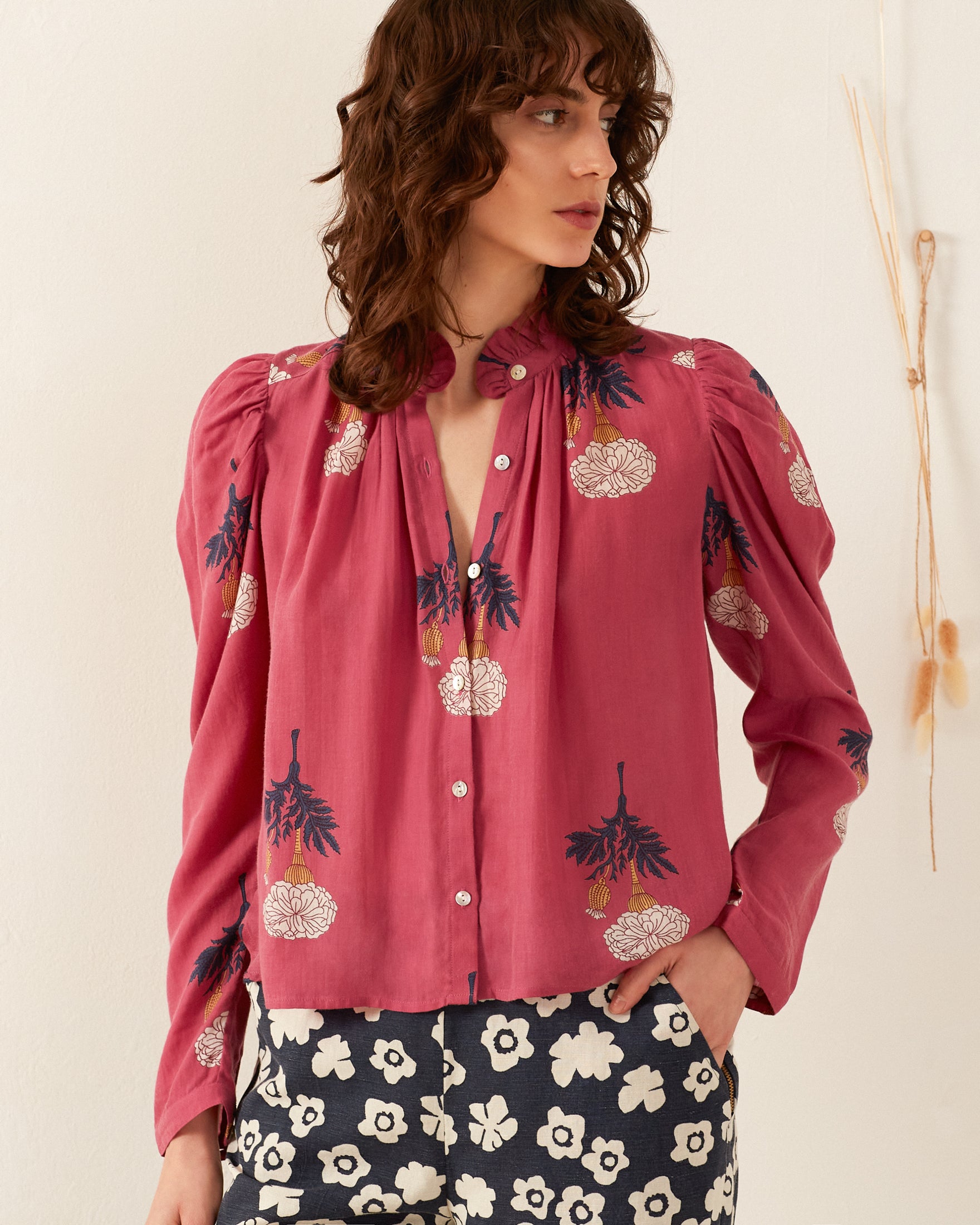 A person with wavy brown hair exudes a vintage feel while wearing the Arte Hibiscus Rose Shirt, a pink button-up blouse adorned with floral patterns, paired with navy blue floral-patterned pants. They're posing in front of a white wall with minimal decor.