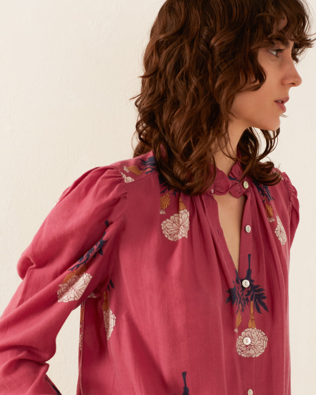 A person with wavy brown hair is modeling the Arte Hibiscus Rose Shirt, a pink blouse adorned with floral patterns and a vintage vibe. The shirt features long sleeves with puffed shoulders and a high neck, complete with buttons down the front. They are depicted in profile against a light background.