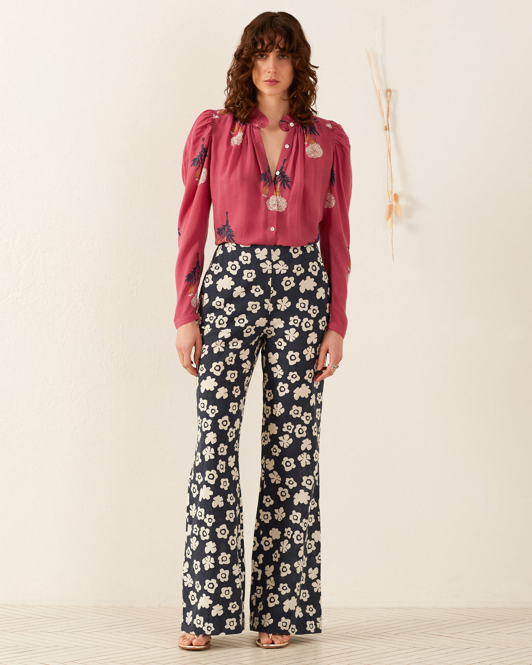 Wearing the Arte Hibiscus Rose Shirt paired with high-waisted, wide-leg pants adorned with a white flower pattern, a person exudes a vintage vibe against a plain light background. Dried flowers are decoratively hung on the wall beside them.