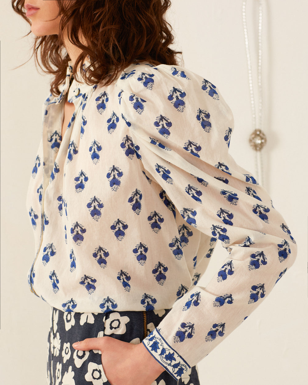 A woman is wearing the Artemis Indigo Bhutti shirt, featuring an off-white design with blue floral patterns, paired with a dark skirt decorated with large white flowers. Her shoulder-length, wavy hair enhances the elegance of her hand-block printed ensemble. The image captures her from shoulders to waist, slightly turned to the side, adding sophistication to her look.