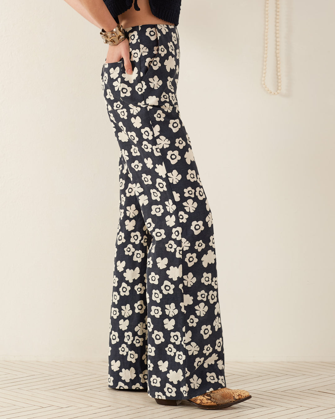 A person wearing the Charlie Deep Sea Lotus Pant, featuring high-waisted, '70s-style floral patterns and bell-bottoms, stands sideways. Their hand rests in the pocket, elegantly complemented by a bracelet. The backdrop is neutral-colored.