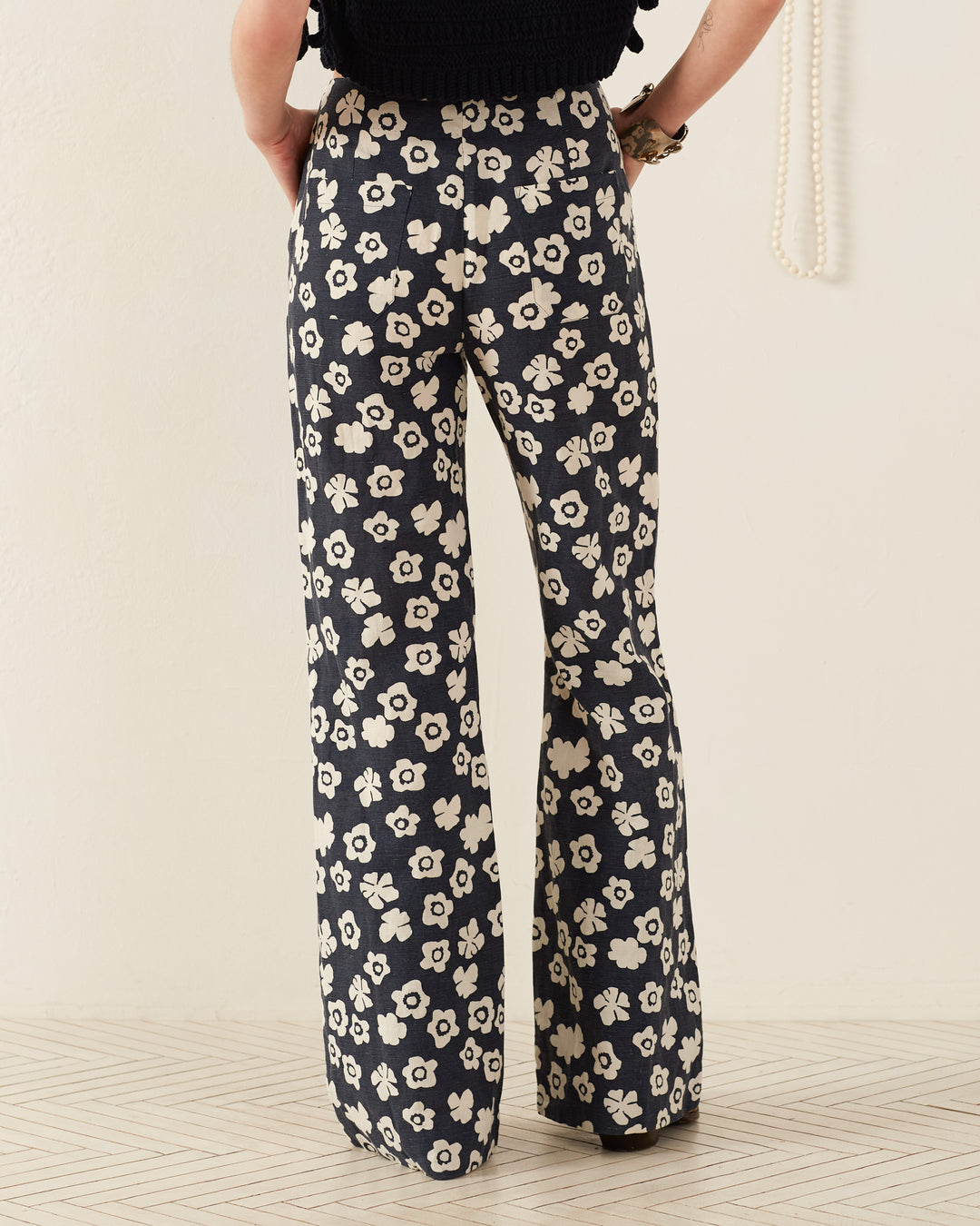 A person wearing the Charlie Deep Sea Lotus Pant, which showcases a black and white floral pattern in a '70s-style wide-leg design, stands facing a light-colored wall. A hand holding a string of pearls is slightly visible to the right. The floor displays a subtle geometric pattern.