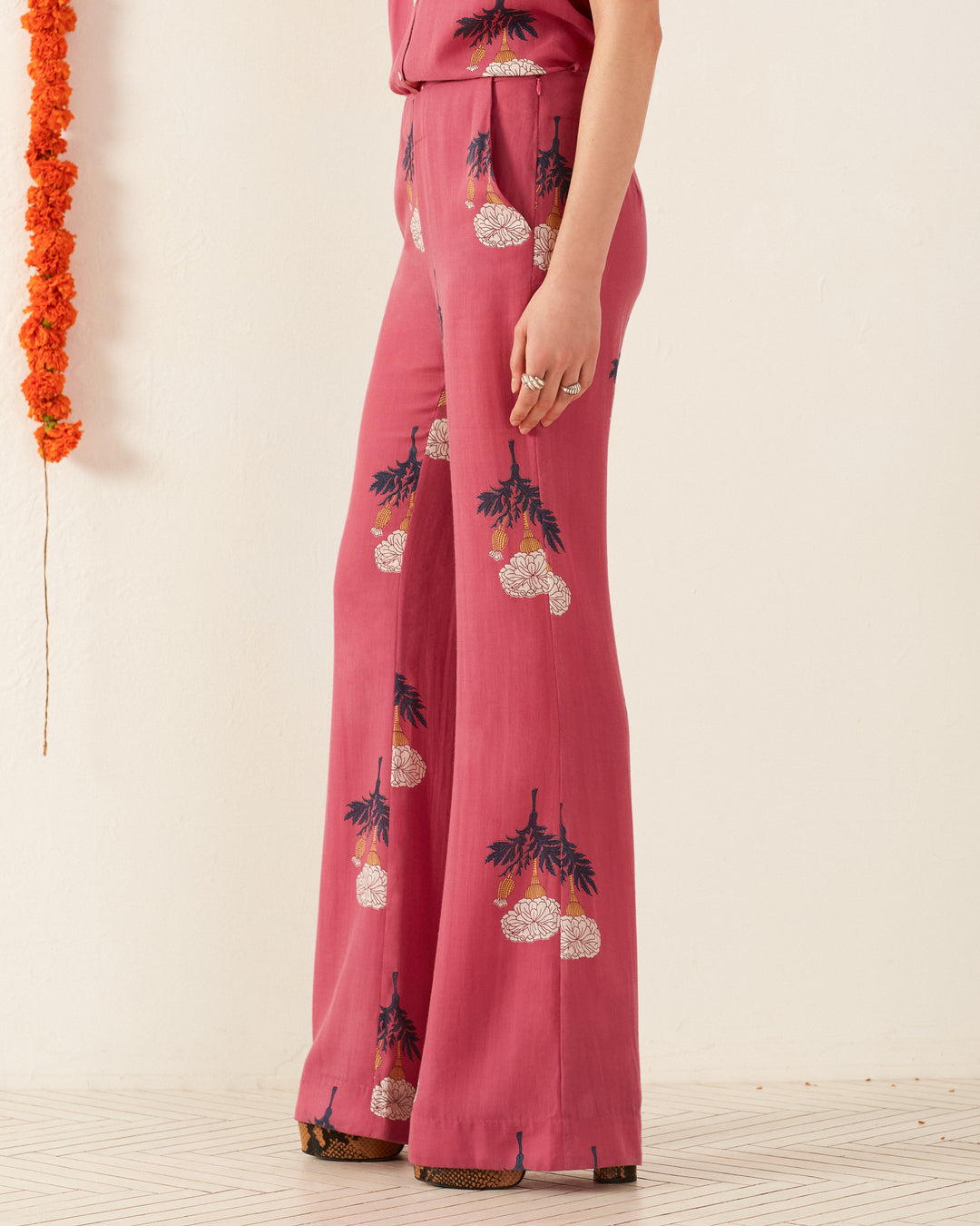 A person wearing the Charlie Hibiscus Rose Pant, featuring high-waisted, wide-leg design with a retro floral pink pattern, stands against a cream background, exuding a vintage vibe. Orange flowers hang from the wall on the left.