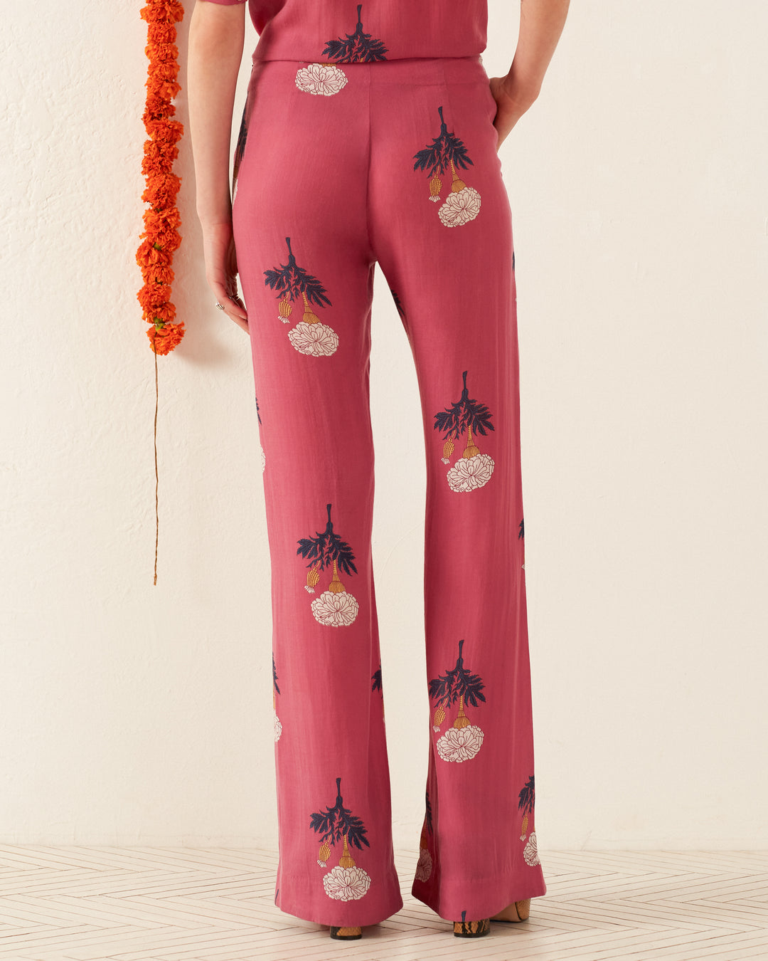 A person wearing the Charlie Hibiscus Rose Pant is captured from behind, adorned with retro floral designs in pink. To the left, orange flowers contribute to a vintage ambiance, while the off-white wall and light-colored floor set a subtle backdrop.