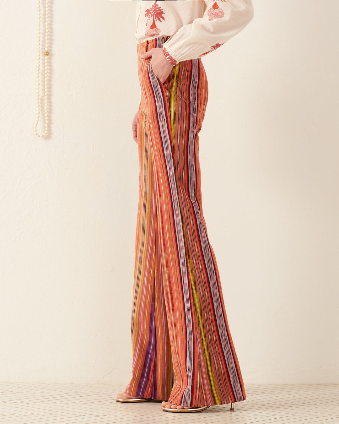 A side view showcases a person wearing the vibrant, retro Charlie Melon Stripe Ikat Pant paired with a white blouse featuring red floral embroidery. They stand against a light-colored wall, with pearls draping in the background while their high-heeled shoes elegantly complete the '70s-inspired look.