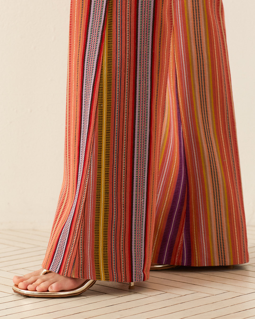 A person wearing the vibrant Charlie Melon Stripe Ikat Pant stands on a light wooden floor, showcasing their bare foot and a pair of minimalist sandals. The pants feature colorful '70s-style vertical stripes in shades of red, orange, pink, yellow, purple, and blue.