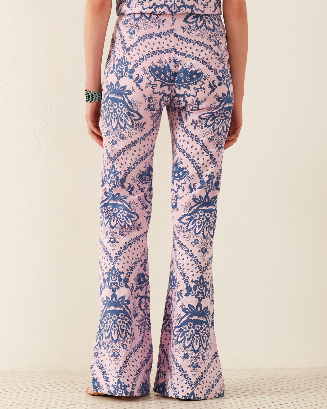 A back view of an individual wearing the Charlie Oasis Bandana Pant, featuring retro flared legs adorned with pink and blue patterns. The complex floral and abstract designs echo a Guatemalan ikat aesthetic. They are standing on a light-colored floor against a plain wall.