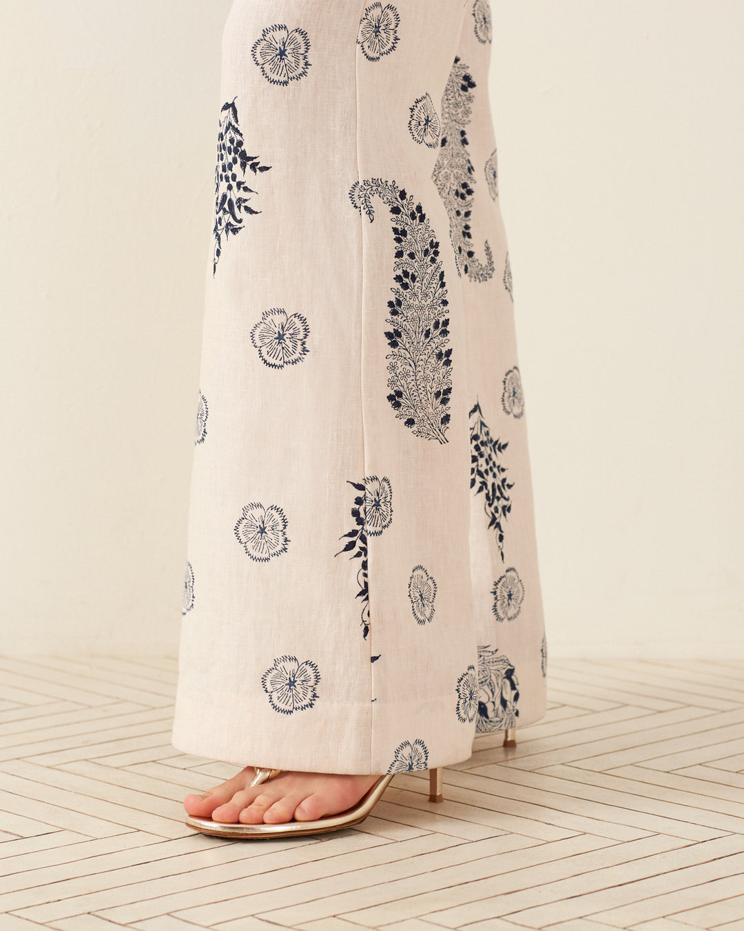 A person wearing the Charlie Porcelain Linen Pant, featuring '70s-style wide-legged design with navy floral and paisley prints, stands on a light-colored, patterned floor. The hand-loomed cotton fabric of the beige heeled sandals perfectly complements the minimalist and stylish scene.