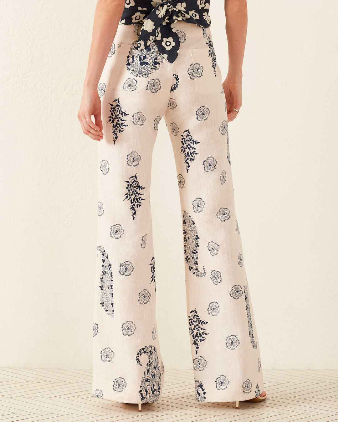 A back view of a person is shown wearing the Charlie Porcelain Linen Pant, featuring a wide-leg, high-waisted design with a '70s-style floral motif in cream. These flowy pants are paired with a matching floral top, both crafted from hand-loomed cotton. The scene is set against patterned flooring and a plain, light-colored wall background.