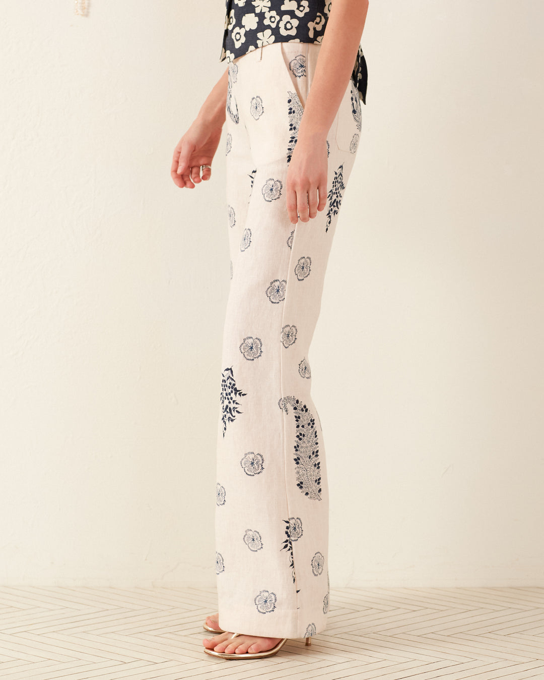 A person wearing cream-colored Charlie Porcelain Linen Pants with a '70s-style floral motif and a matching top stands sideways, highlighting the design of the pants. The wooden floor contrasts with the plain wall as they remain barefoot.