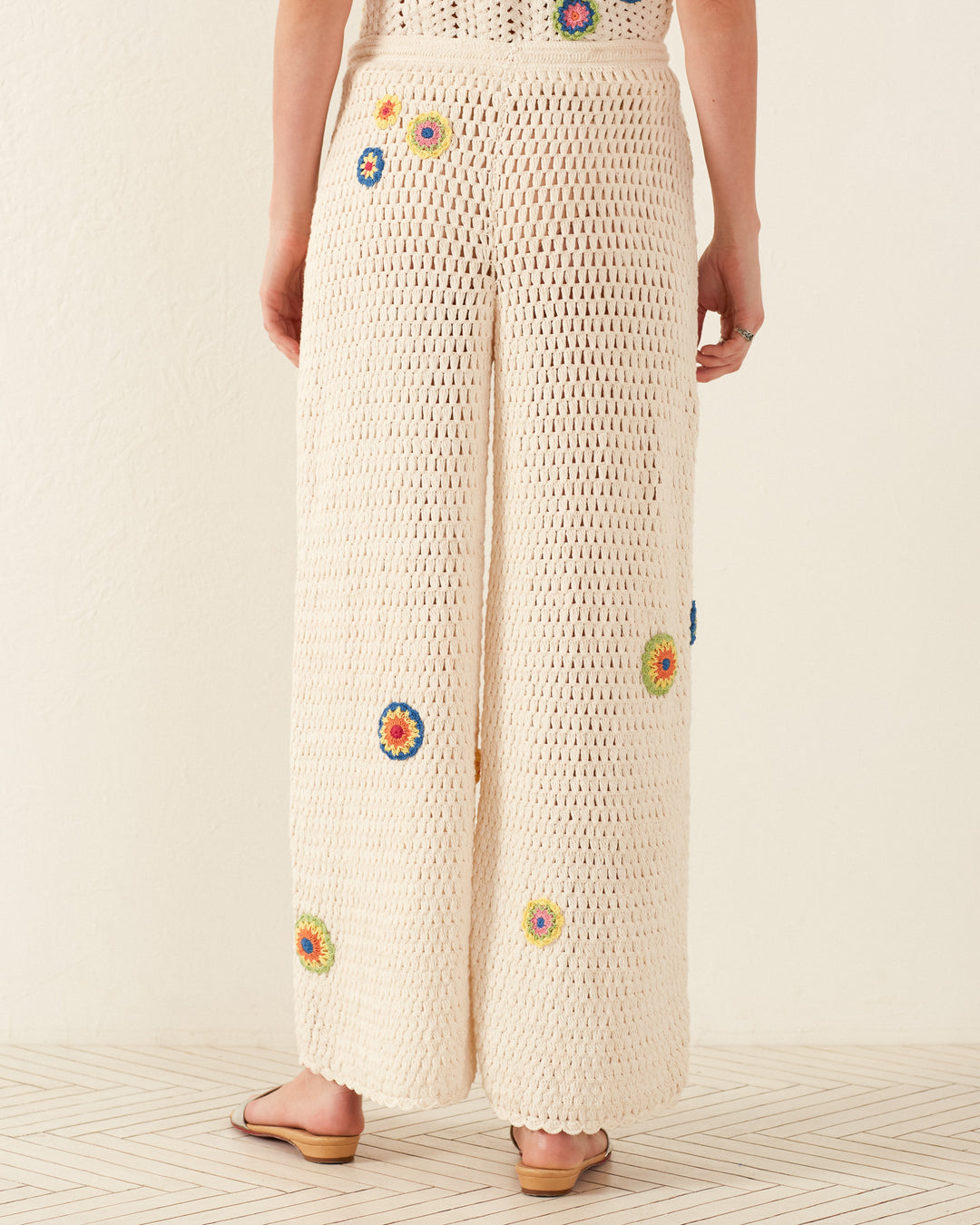 A person wearing the Elia Pearl Flower Crochet Pant, adorned with colorful floral patterns, and a Victorian camisole stands on a patterned floor. The loose-fitting pants pair effortlessly with flat sandals, while the off-white background accentuates the vintage style.