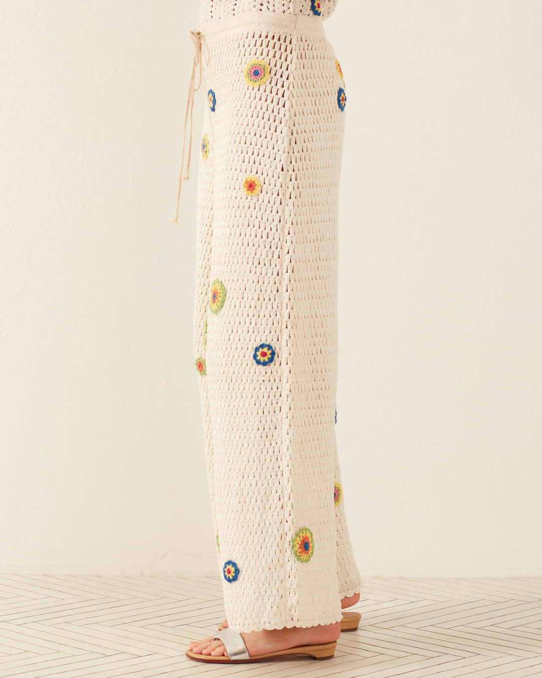 A person is wearing the Elia Pearl Flower Crochet Pants, which are cream-colored with colorful floral patterns and a drawstring waist. The pants have wide legs and are paired with a Victorian camisole. They stand sideways on a wooden floor against a plain, light-colored wall.