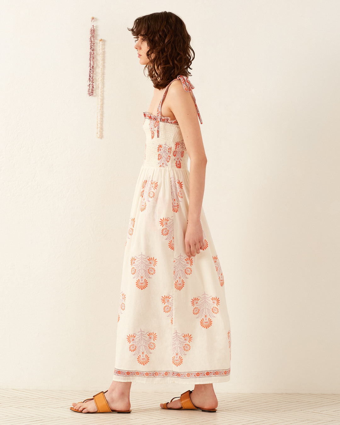 A woman stands in profile wearing the Kaia Papaya Aster Dress, a sleeveless white maxi dress adorned with orange floral patterns. She has curly hair and wears yellow sandals. Behind her, a beaded decoration hangs on the wall, reminiscent of a Western jacket with its intricate details.