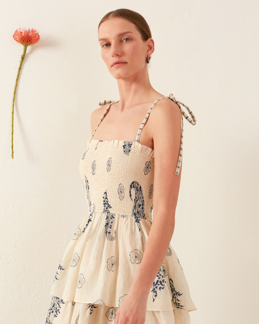 A woman wearing the cream-colored Kaia Porcelain Tier Dress, featuring a delicate floral print and thin, tied straps, stands against a plain white wall. A single flower with a long stem is attached to the wall beside her.