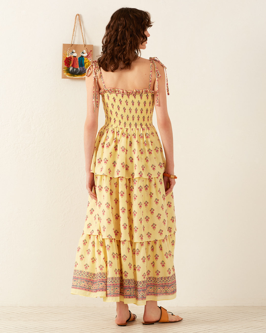 A woman wearing the Kaia Primrose Tier Dress, which features flowing cotton voile and floral patterns, stands with her back to the camera. She pairs it with sandals and leans slightly to the left. In the background is a decorative wall hanging adorned with colorful figures.