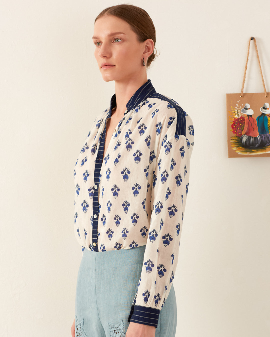 A person with tied-back hair wears the Kiki Indigo Bhutti Shirt, featuring block-printed long sleeves and blue designs, paired with light blue pants. They stand in a minimalist room adorned by a small decorative painting on the wall.