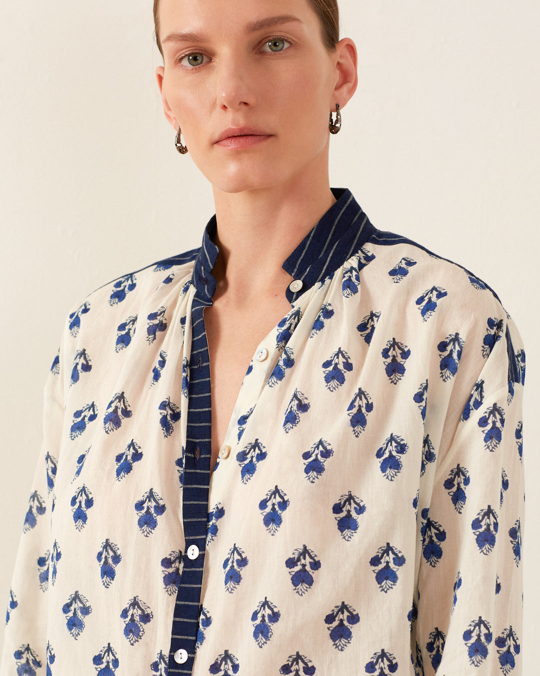 A person with short hair is wearing the Kiki Indigo Bhutti Shirt, characterized by its open collar and long sleeves adorned with block-printed blue floral patterns on a white background. The shirt features striped trim along the collar and down the front, perfectly complemented by hoop earrings for a stylish contrast to a summer dress look.