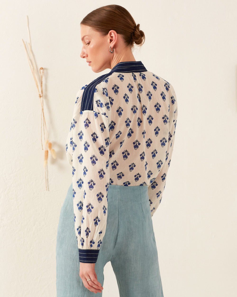 A woman with braided hair stands facing away, showcasing the Kiki Indigo Bhutti Shirt paired with light blue pants. The shirt's subtle block-printed design and navy accents add charm as she stands in front of a neutral wall with minimal decor.