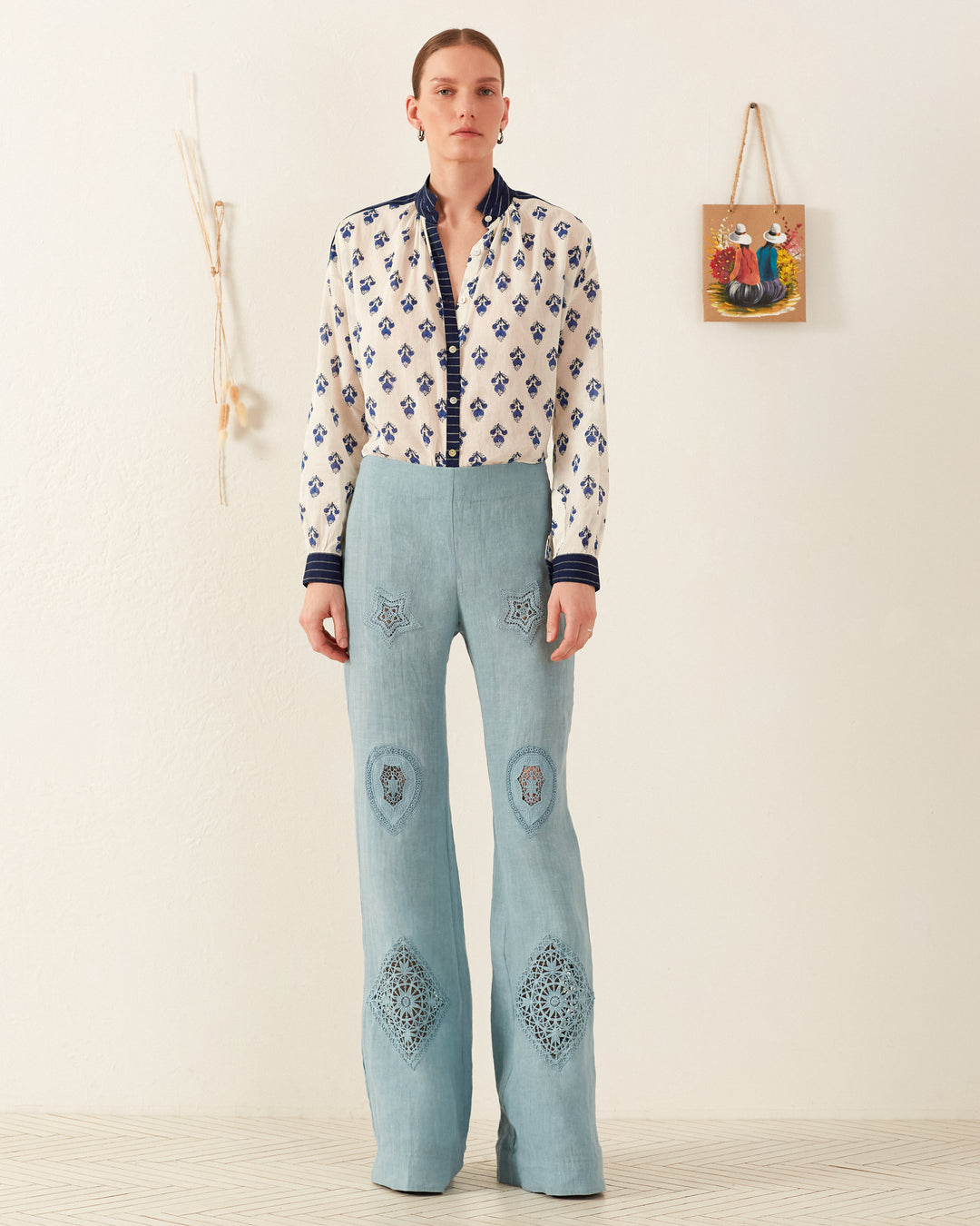 A person stands against a white wall wearing light blue high-waisted pants with intricate block-printed designs, paired with the Kiki Indigo Bhutti Shirt. Their hair is pulled back. The wall has minimal decor, including a small painting and a hanging ornament.