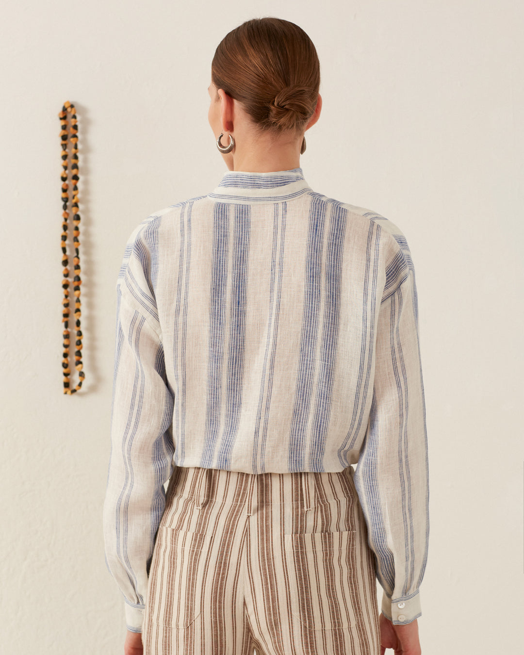 A person with brown hair in a bun is facing away, wearing a Kiki Pacific Star Shirt featuring a light blue and white floral print, paired with brown striped pants. The cotton voile shirt complements the beaded necklace hanging on a white wall to the left.