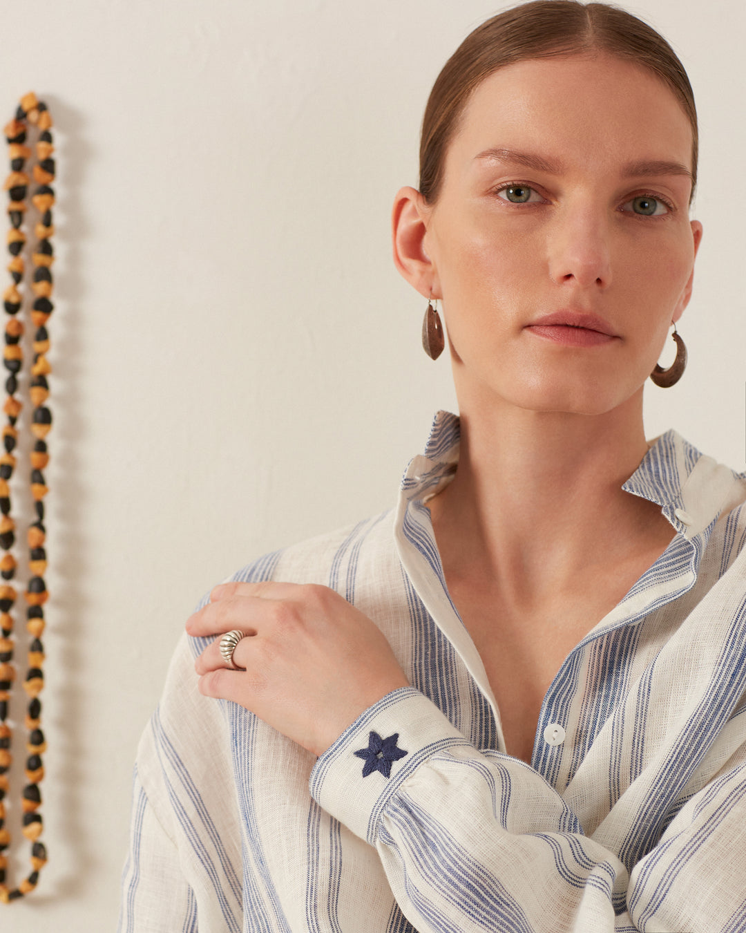 A person with light-colored hair is wearing the Kiki Pacific Star Shirt, a hand-block printed, striped button-up shirt featuring a star on the cuff. They are accessorized with hoop earrings and a ring. In the background, a beaded necklace hangs on the wall.