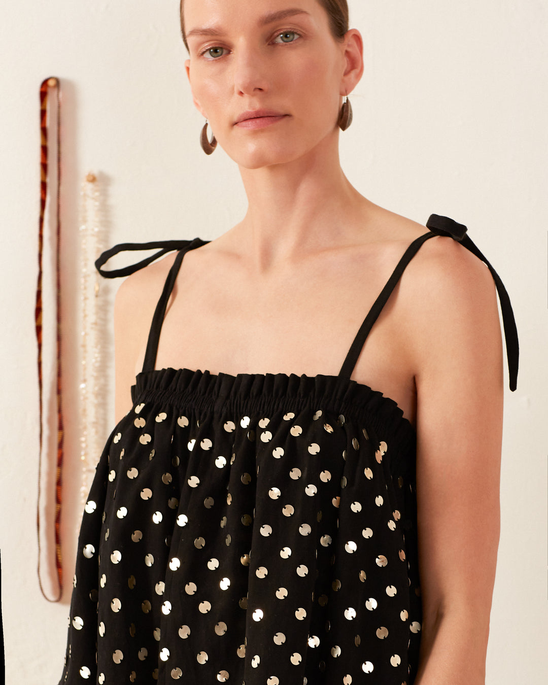 A person wearing the Lila Night Sky Coin Top, featuring a black design with silver polka dots and tied shoulder straps, stands against a white background. Behind them, hanging decorations adorn the wall.