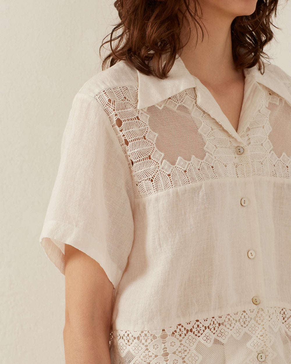 A person wearing the Stevie Picnic Eyelet Shirt, a short-sleeved, button-up white blouse featuring intricate lace detailing on the shoulders and upper chest. Made from soft, hand-loomed cotton, it evokes a gentle summer vibe as the person stands against a light-colored background.