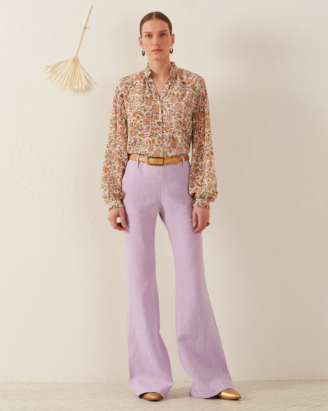 A person stands against an off-white background, wearing the Poet Garden Party Silk Blouse and lavender wide-legged pants paired with gold shoes. They have short hair and are accessorized with hoop earrings and a belt. A decorative fan is on the wall behind them.