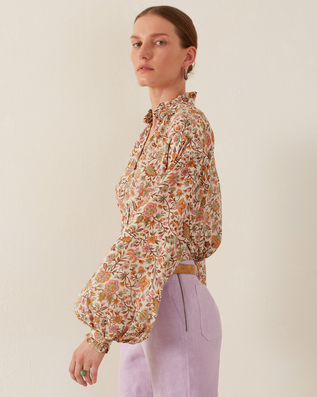 A person with short hair wearing the Poet Garden Party Silk Blouse, adorned with block-printed pink and orange flowers and green leaves, along with lilac pants, stands against a plain background. The blouse features voluminous sleeves as they gaze to the side.