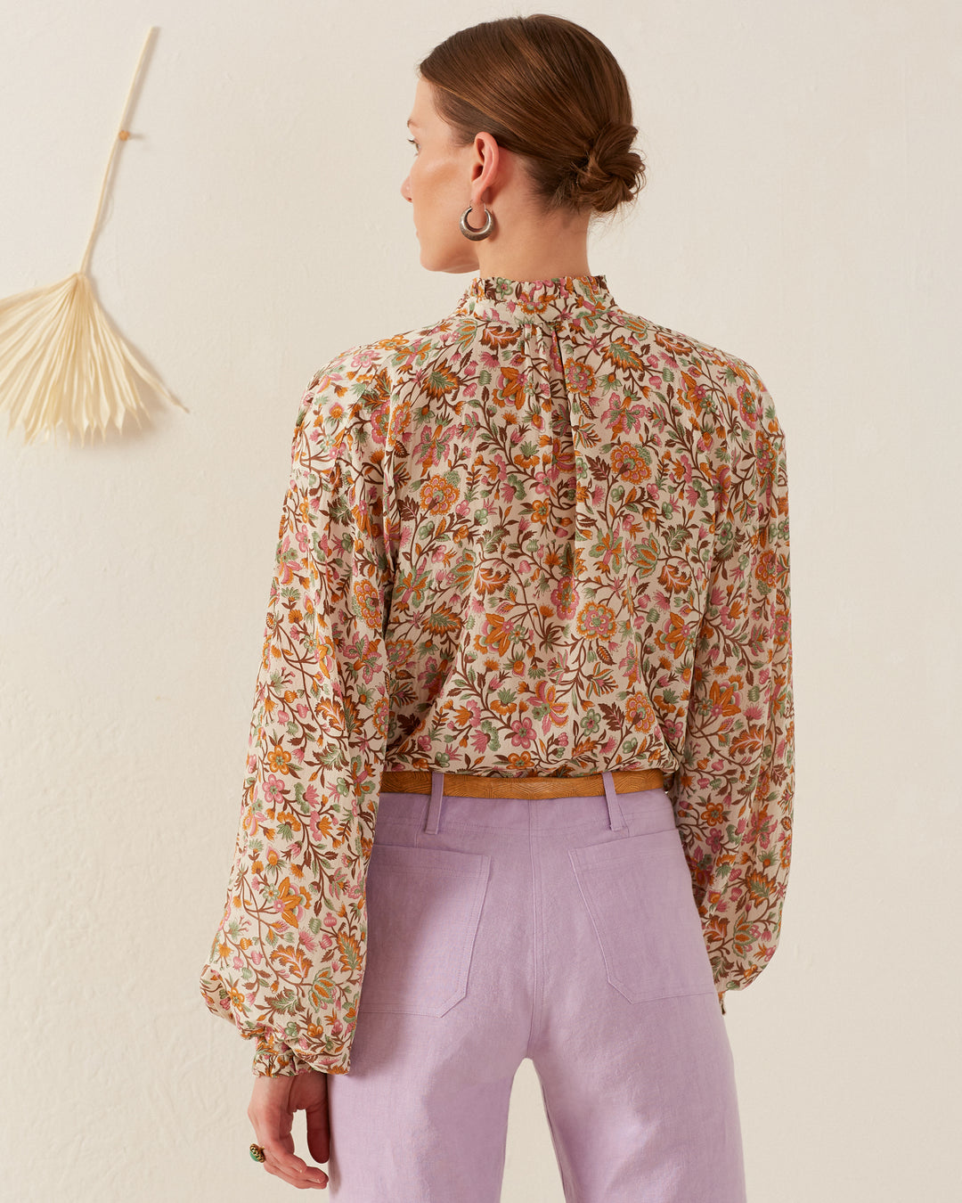 A person stands against a light-colored wall, facing away. They wear the Poet Garden Party Silk Blouse, paired with lavender pants that give off a vintage pajama style vibe. A dried palm leaf decorates the wall on the left.