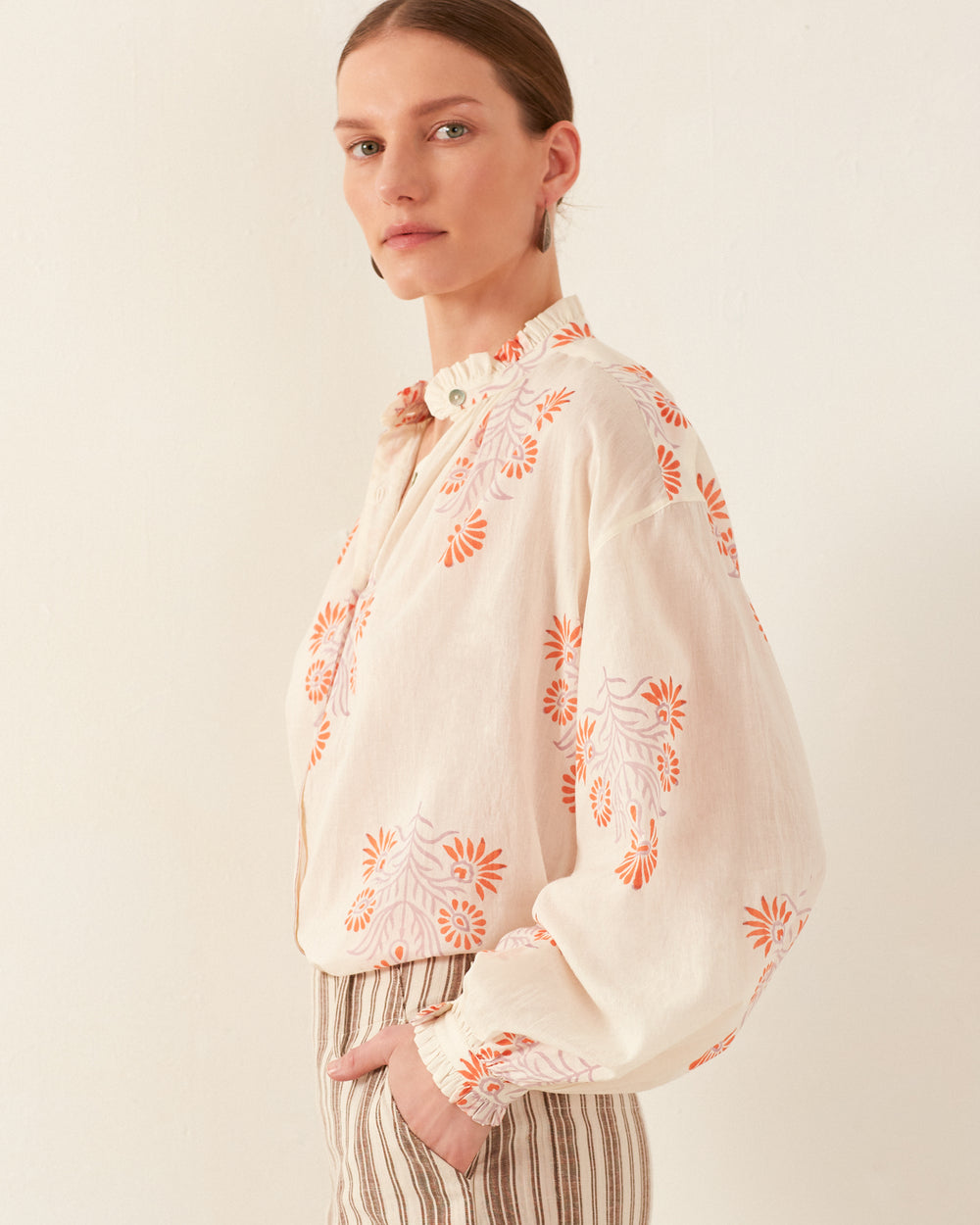 A person wearing the Poet Papaya Aster Blouse, a cream-colored, vintage-inspired top adorned with orange floral embroidery, pairs it with striped pants against a plain background. The elegant silk chiffon shirt features long sleeves, and their hair is neatly pulled back.