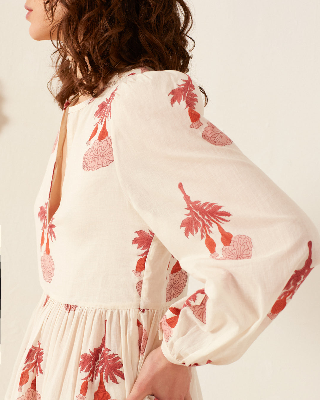 A woman wearing the Silas Tea Rose Dress, a cream outfit adorned with red floral patterns, crafted from soft cotton voile. The dress showcases long sleeves and a V-neckline, beautifully complementing her curly brown hair as she stands in profile against a plain white background.