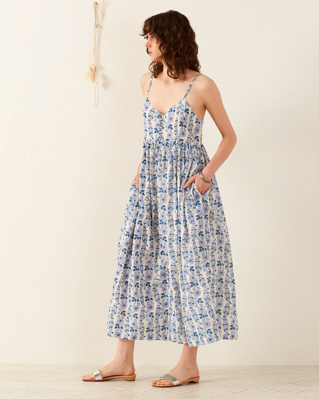 A person with curly hair is wearing the Skylar Iris Ivy Dress, a sleeveless, hand-block printed cotton voile piece featuring blue and white floral patterns. With hands casually in pockets, they stand slightly angled to create a striking pose against a minimalist backdrop of light-colored flooring.