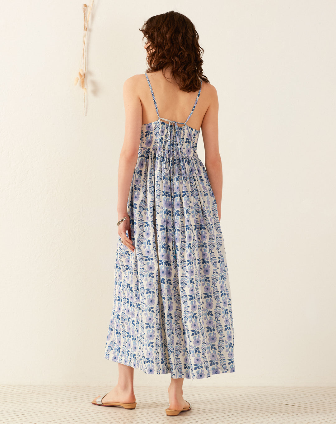 A person with long, wavy brown hair stands facing away, wearing the Skylar Iris Ivy Dress featuring a hand-block printed floral design in blue and white patterns with thin straps. They are in a minimalistic white room decorated with cotton voile curtains and a small decorative item on the wall.