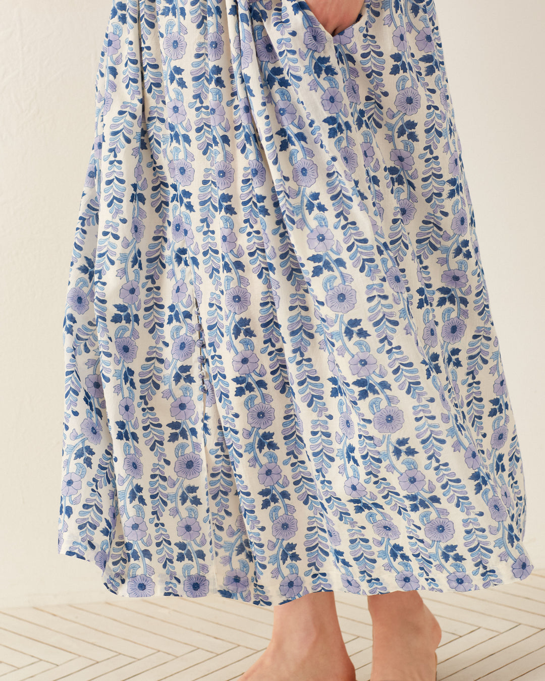 A person wearing the Skylar Iris Ivy Dress, made from hand-block printed cotton voile showcasing a blue floral pattern, stands barefoot on a light-colored, textured floor. The background is a plain off-white wall.