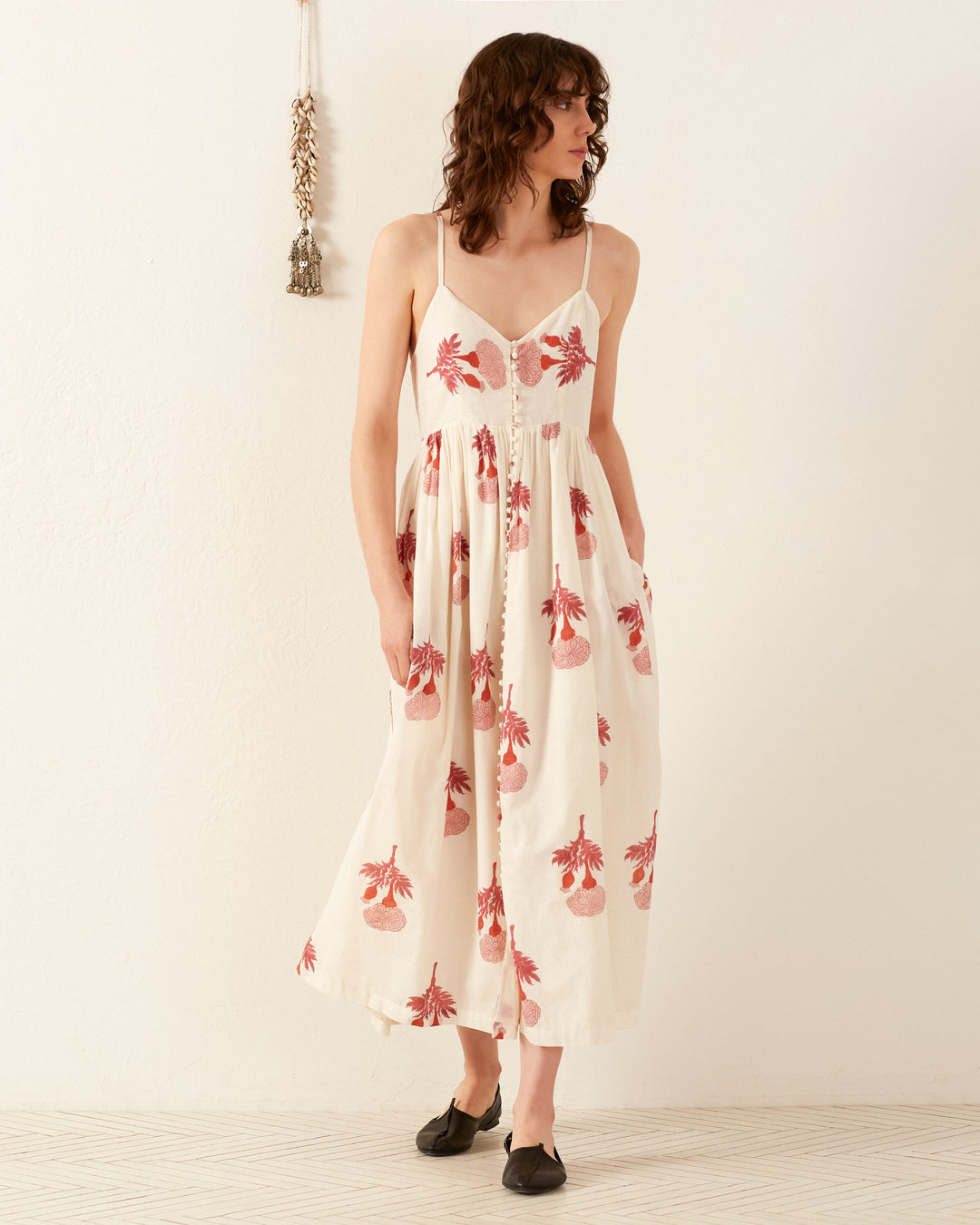 A person stands against a white wall wearing the Skylar Tea Rose Dress, which is a cream, sleeveless summer dress with red floral patterns. They have curly, shoulder-length hair and are barefoot on a tiled floor. A small decorative item hangs on the wall.