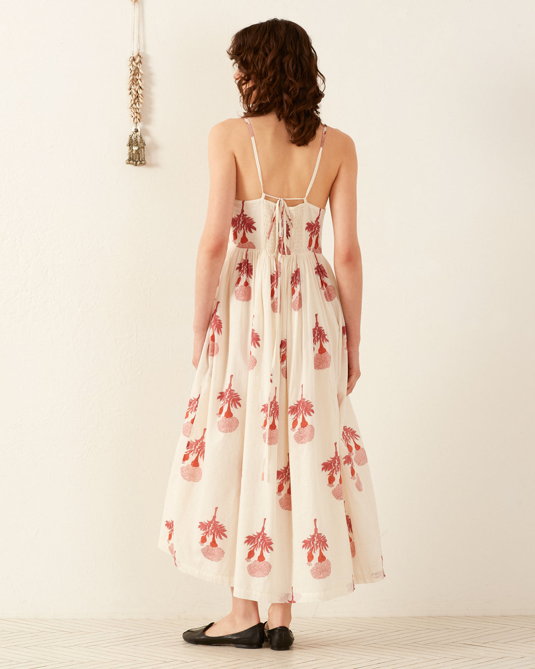 A person with wavy hair stands facing a white wall, wearing the Skylar Tea Rose Dress, a long summer dress adorned with red floral patterns. The dress features thin straps and a lace-up back, exuding charm. To the left, a decorative wall hanging complements the scene.