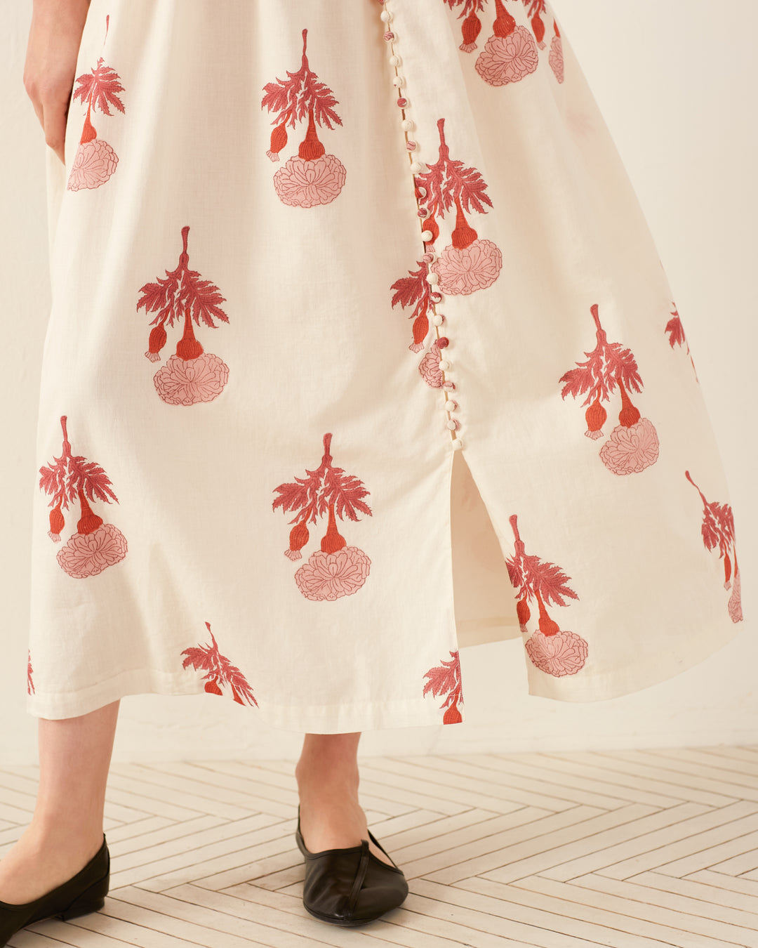 A person wearing the Skylar Tea Rose Dress, which is cream-colored with a red floral pattern and hand block-printed, stands on a herringbone-patterned floor. The dress features a row of buttons down the side and is paired with black slip-on shoes.