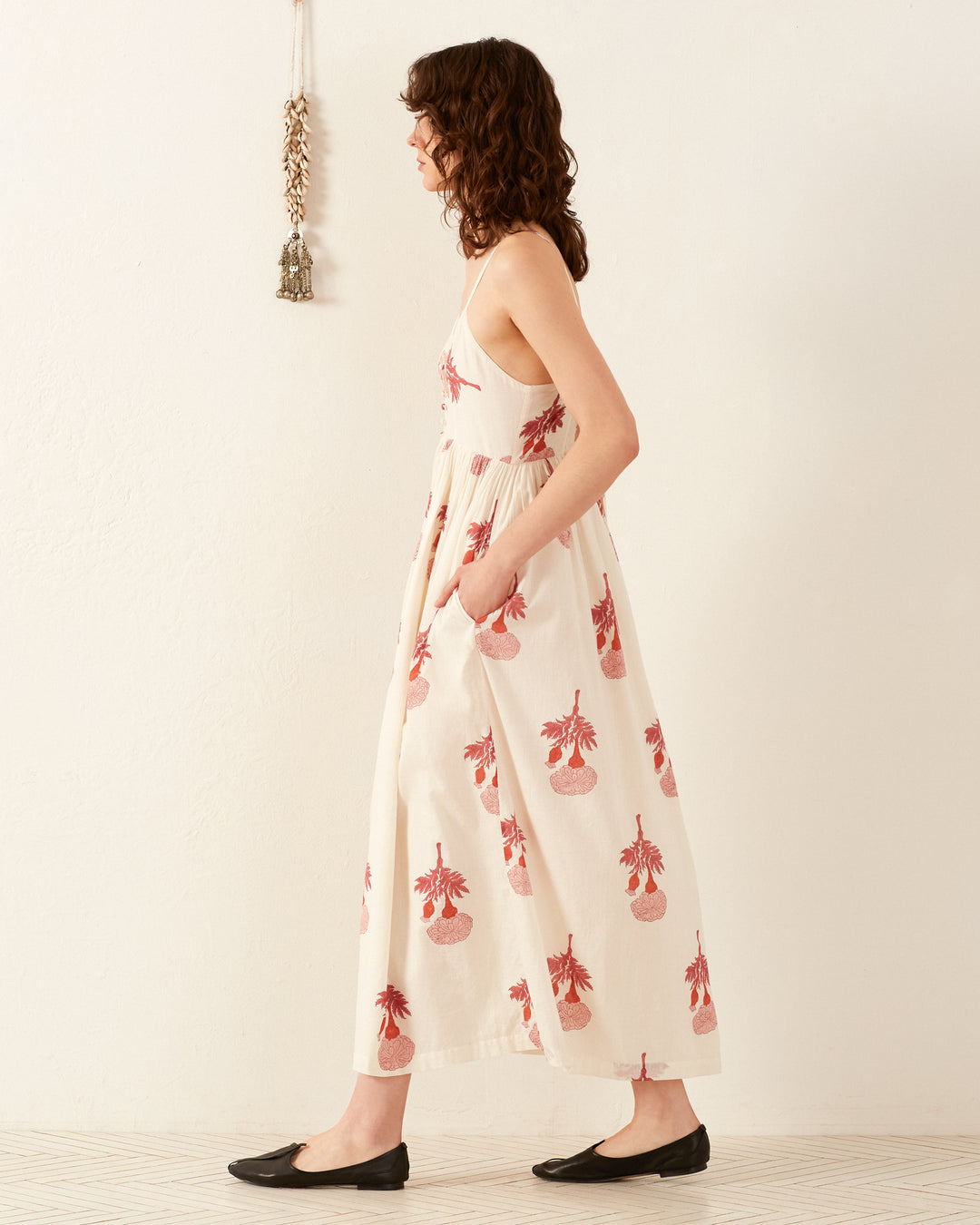 A woman in the Skylar Tea Rose Dress, featuring a white sleeveless design with red floral patterns, stands sideways against a plain wall, embodying the essence of a breezy summer dress. Her brown curly hair complements her look, as she wears black flats and is accompanied by a small decorative hanging on the wall behind her.