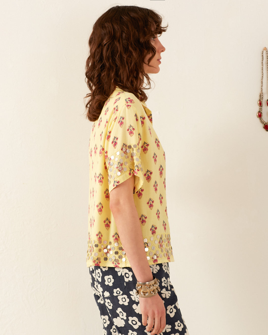 A woman with curly brown hair stands in profile against a plain background, wearing a Stevie Primrose Coin Shirt adorned with red floral patterns and shiny embellishments. Her outfit is completed with black and white floral pants, accessorized by a bracelet.