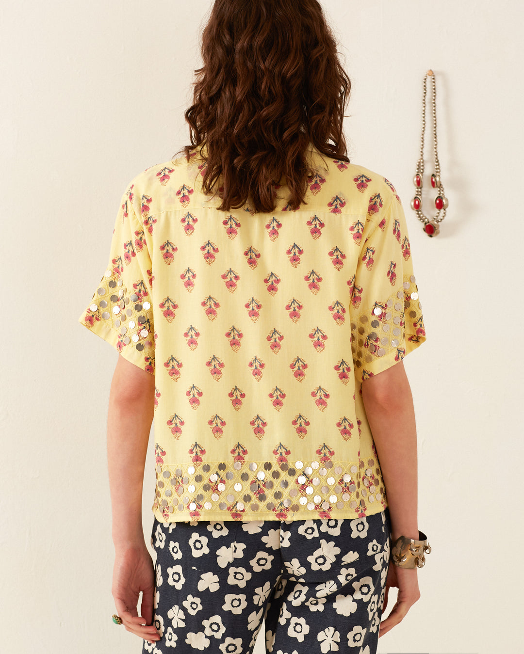 The image depicts the back view of a person dressed in the Stevie Primrose Coin Shirt, which is yellow with a vintage Hawaiian pattern featuring red floral designs and reflective circles near the hem. The person pairs it with navy and white floral pants. A necklace is visible hanging on the wall behind them.