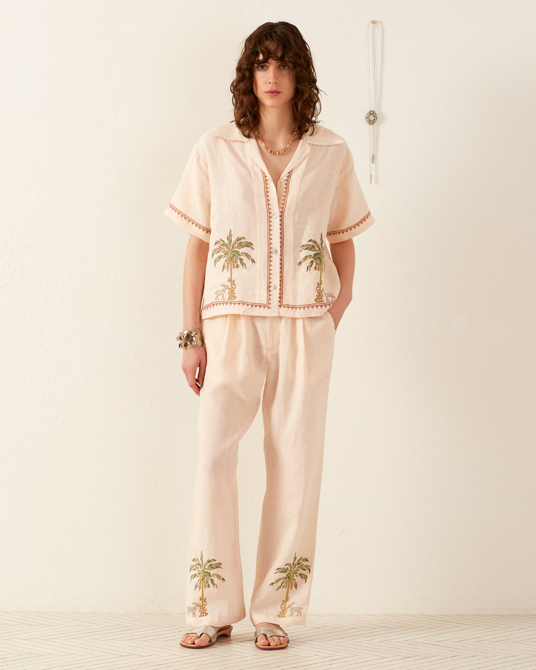 A person with wavy hair stands against a plain white background wearing the Stevie Sanded Palm Shirt, a light beige cotton piece featuring palm tree embroidery. They are accessorized with a necklace and bracelets and are wearing sandals.