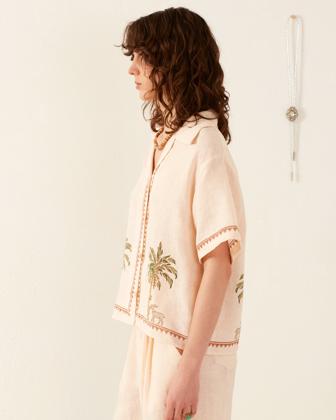 A woman with curly brown hair stands sideways against a white background, wearing the Stevie Sanded Palm Shirt, a vintage Hawaiian top featuring palm tree designs. A long necklace hangs on the wall beside her.