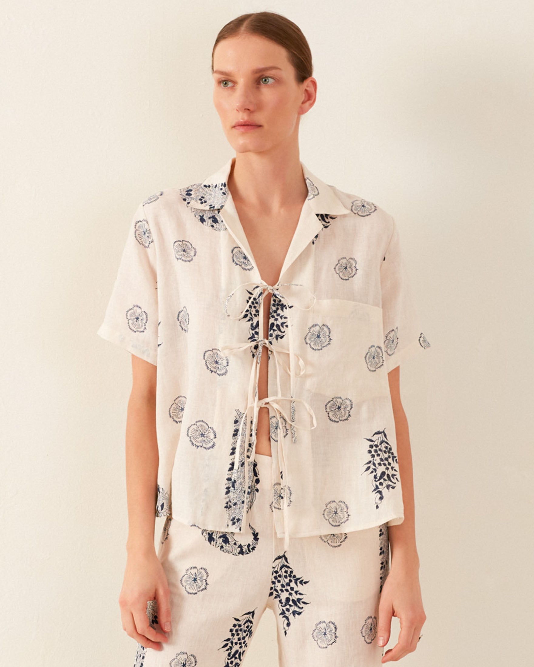 A person wearing the Styla Porcelain Linen Shirt with a floral pattern stands against a plain background. The linen shirt features a lace-up front reminiscent of a vintage Hawaiian style, and the trousers complement it with a matching design. The overall look is casual and summery.