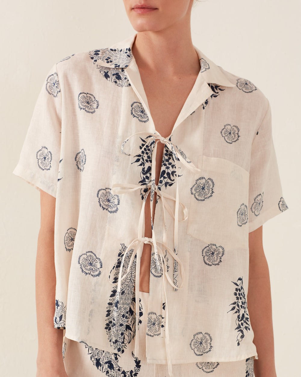 A person is wearing the Styla Porcelain Linen Shirt, a light top with short sleeves and front tie closures. It features navy blue floral designs on a cream background reminiscent of a vintage Hawaiian shirt, set against a plain, neutral backdrop that allows the charming design to stand out.