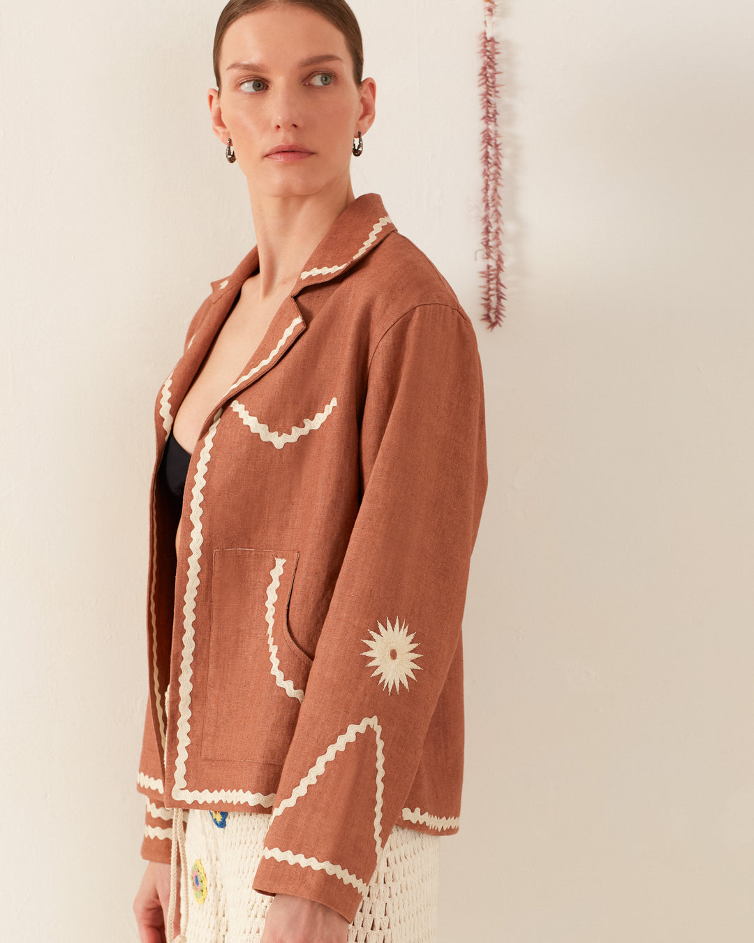 A person stands against a beige wall wearing the Western Clove Ric Rac Jacket, featuring mother-of-pearl buttons and white embroidered trim, with a black top peeking through. They also wear a white textured skirt. A thin red decoration hangs on the wall behind, casting subtle charm in the retro ambiance.